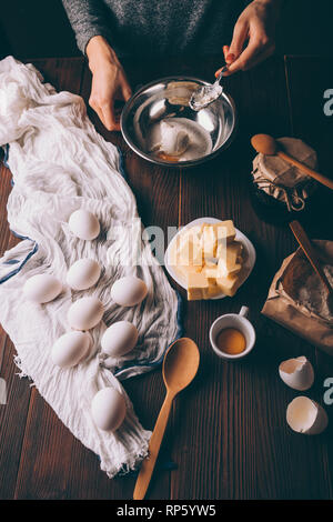 Ansicht von oben junge Frau Kochen Teig für Kreis mischen Saure Sahne, Zucker und Eigelb in Metall Schüssel Neben Eiern, gewürfelte Butter und Mehl auf braunem Holz- Stockfoto