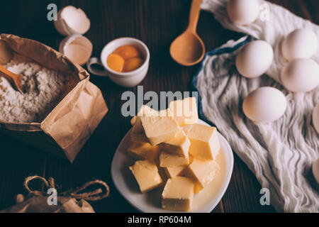 Rustikale Zusammensetzung der Zutaten zum backen hausgemachte Kuchen. Blick von oben auf die gewürfelte Butter, Papier Sack Mehl, rohe Eier, Eigelb und Muscheln auf braunem Holz- ta Stockfoto