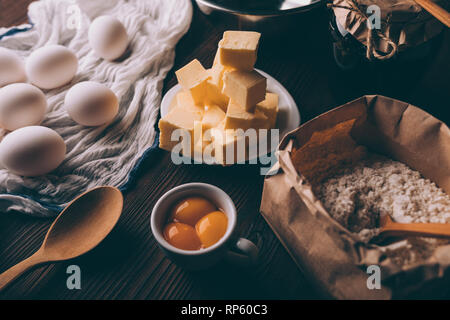 Nahaufnahme der Zutaten zum Kochen Kuchen auf dunklen Holztisch. Gewürfelte Butter, rohe Eier, Eigelb, Mehl, bereit für die hausgemachten Backwaren. Stockfoto