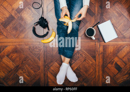 Blick von oben der jungen Frau sitzt auf Holzboden zu Hause Banane schälen in der Nähe von Kaffee, Ohrhörer und leere Notepad. Stockfoto