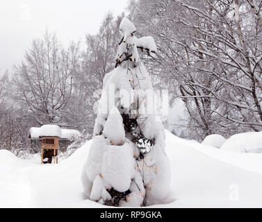 Weihnachtsbaum bedeckt von Schnee und gebeugt unter der Last. Winter und ein Vogel Automaten im Hintergrund. Stockfoto