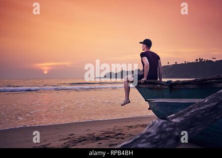 Junge Mann sitzt auf einem Boot, bei Sonnenaufgang. Kontemplation am idyllischen Sandstrand in der Nähe von Tangalle in Sri Lanka. Stockfoto