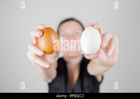 Kaukasische Frau mit schwarzen T-Shirt Holding und zwei Eier zwischen den Fingern, verschwommene Gesicht auf einem weißen Hintergrund. Stockfoto