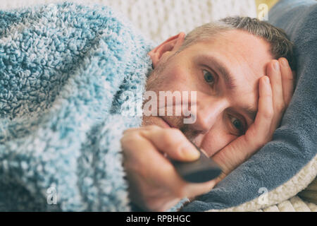 Ältere Menschen, die unter Depressionen leiden, liegt leider mit einer Decke auf seinem Bett und Fernsehen abgedeckt Stockfoto