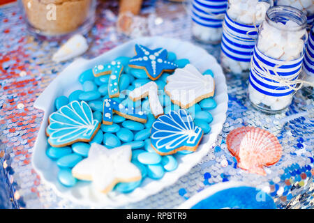 Weiße Muschel Platte voller Pralinen mit blauer Zuckerglasur, Cookies wie Muscheln, Sterne und Nummer Sieben und viele kleine Gläser mit Marshmallow auf t Stockfoto