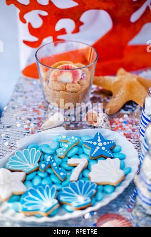 Weiße Muschel Platte voller Pralinen mit blauer Zuckerglasur, Cookies wie Muscheln, Sterne und Nummer Sieben und viele kleine Gläser mit Marshmallow auf t Stockfoto