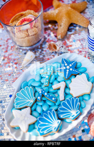 Weiße Muschel Platte voller Pralinen mit blauer Zuckerglasur, Cookies wie Muscheln, Sterne und Nummer Sieben und viele kleine Gläser mit Marshmallow auf t Stockfoto