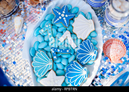 Weiße Muschel Platte voller Pralinen mit blauer Zuckerglasur, Cookies wie Muscheln, Sterne und Nummer Sieben und viele kleine Gläser mit Marshmallow auf t Stockfoto