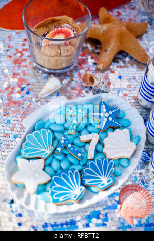 Weiße Muschel Platte voller Pralinen mit blauer Zuckerglasur, Cookies wie Muscheln, Sterne und Nummer Sieben und viele kleine Gläser mit Marshmallow auf t Stockfoto