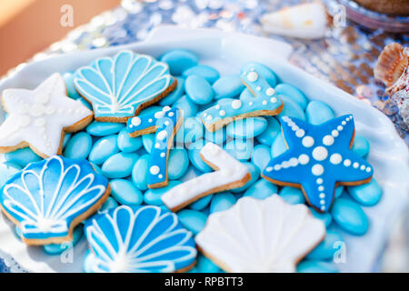 Weiße Muschel Platte voller Pralinen mit blauer Zuckerglasur, Cookies wie Muscheln, Sterne und Nummer Sieben und viele kleine Gläser mit Marshmallow auf t Stockfoto