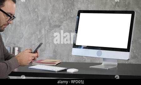Blick auf den Bildschirm des Computers. Junger Mann SMS am Telefon mit einem Computer auf dem Schreibtisch. Weißes Display. Professionelle Aufnahme in 4K-Auflösung. 007. Yo Stockfoto