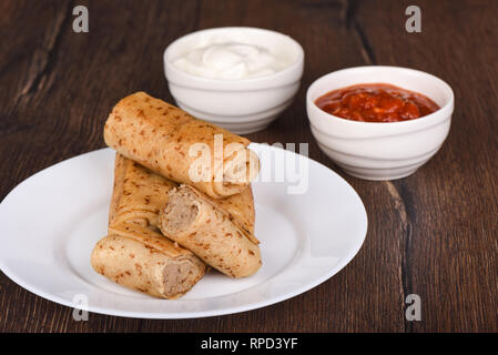 Pfannkuchen Brötchen mit Fleisch füllen, serviert mit saurer Sahne, Ketchup Sauce. Die traditionellen russischen Speisen auf Holz- Hintergrund Stockfoto