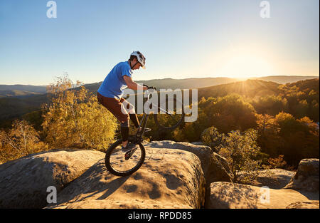 Radprofi reiten auf Hinterrad auf Versuch Fahrrad. Sportler Reiter balancing am Rande des großen Felsen auf dem Gipfel des Berges bei Sonnenuntergang. Konzept der Extreme Sport aktiven Lebensstil Stockfoto