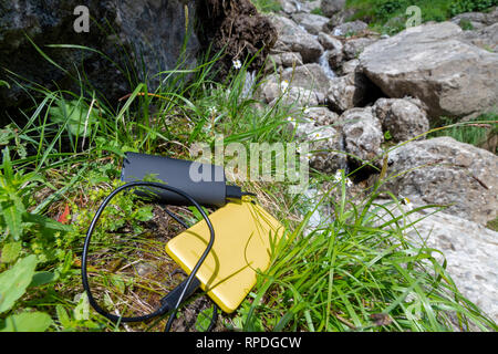 Gelbe Smartphone aus einem schwarzen powerbank externe Batterie aufgeladen, auf Gras, in der Nähe von Felsen, oben in den Bergen. In Verbindung bleiben beim Wandern con Stockfoto