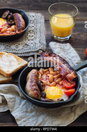 Gebratene Eier, Würstchen, Speck, Bohnen und Champignons in Iron Skillet, Toast, Orangensaft, Butter auf rustikalen Holzmöbeln Hintergrund. Englisches Frühstück. Stockfoto