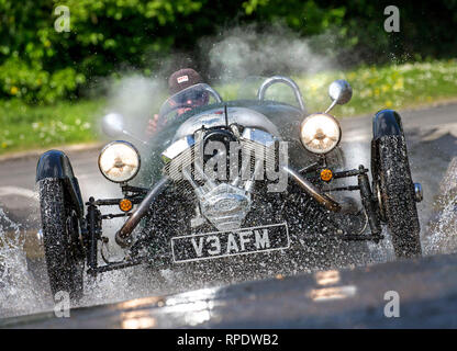 Morgan 3-wheeler Spritzwasser durch eine Furt an Geschwindigkeit mit Scheinwerfern und Reflexionen in der nassen Straße. Model Release Form unterzeichnet. Stockfoto