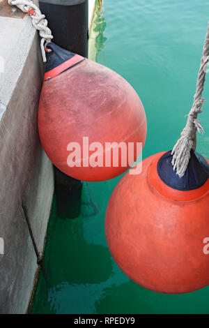 Close-up zwei roten Kunststoff dock festmachen Fender, die auf die Seile in der Nähe von Beton- und auf dem Wasser im Hintergrund an der Wand hängen. Stockfoto