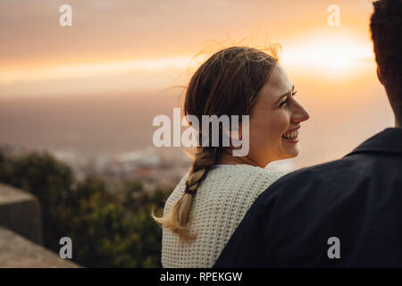 Fröhliche junge Frau sitzt mit ihrem Freund im Freien. Weiblichen Blick auf ihren Mann und lächelnd gegen Sonnenuntergang. Stockfoto