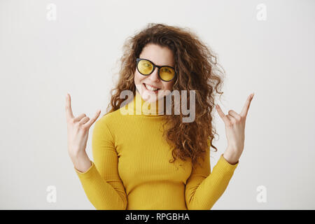 Schön lustig erstaunt Rocker Mädchen mit lockigem Haar und ständigen mit Rock Zeichen und Kamera. studio Shot, auf grauem Hintergrund Stockfoto