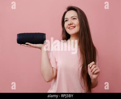 Glückliche junge attraktive Frau auf rosa Hintergrund Halten in der Hand tragbare Lautsprecher, um Musik zu hören Stockfoto