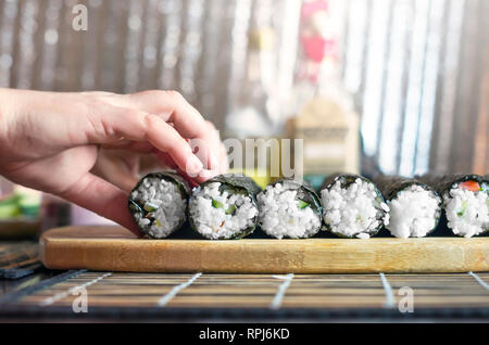 Die Sushi Rollen Workshop. Weibliche Hände Rollen auf dem Holzbrett für die Abdichtung. Küchenutensilien und glänzendes Metall Hintergrund Stockfoto