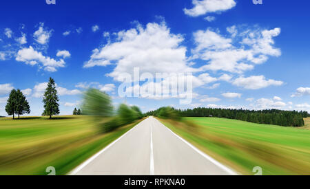 Straße inmitten grüner Felder mit Motion blur Effekt, blauer Himmel und weiße Wolken im Hintergrund Stockfoto