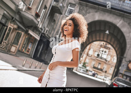 Wahre Schönheit. Portrait von niedlichen Afro-amerikanische Frau im weißen Kleid lachend beim Stehen auf einem sonnigen Straße. Glück Konzept Stockfoto