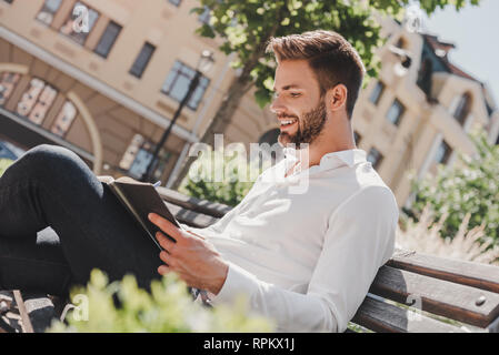 Nahaufnahme von hübscher junger Mann sitzt auf einer Bank im Park und halten seine Routenplaner. Bärtiger Mann in einem weißen Hemd und Jeans durch seine Notizen und lächelnd gekleidet. Sommer, entspannen Stockfoto