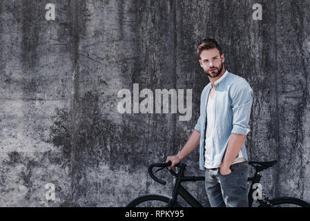 Attraktiven jungen Mann beiläufig in blau Modisches Shirt, Jeans und grauen T-Shirt gekleidet, steht mit schwarzen Fahrrad neben ihm. Er wird zu fahren. Er wirkt nachdenklich, über grauer Hintergrund Stockfoto