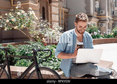 Portrait von gutaussehenden jungen Mann im Park sitzen und seinen Laptop und eine Tasse Kaffee. Braun Bärtigen schwarzhaarige Mann in einem blau gestreiften Hemd und Jeans in seinen Laptop suchen und lächelnd gekleidet. Sommer, entspannen Stockfoto