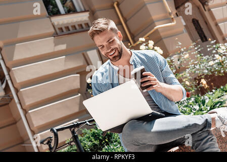 Portrait von gutaussehenden jungen Mann im Park sitzen und seinen Laptop und eine Tasse Kaffee. Braun Bärtigen schwarzhaarige Mann in einem blau gestreiften Hemd und Jeans in seinen Laptop suchen und lächelnd gekleidet. Sommer, entspannen Stockfoto