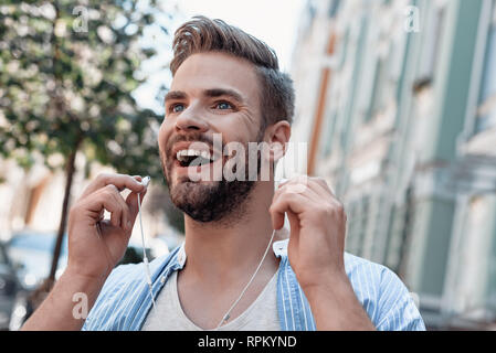 Junge stilvollen Mann mit blauem Hemd und grauen T-Shirt ist zu Fuß im Freien, Musik hören, gerne zur Seite. Er hält seine Kopfhörer, während sich schnell bewegenden Stockfoto