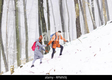 Blonde Kerl trägt orange Jacke hilft, seine Freundin zu der Berg im Winter Forest zu klettern Stockfoto