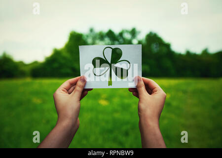 St. Patricks Tag, Glücksbringer, wie menschliche Hände halten ein Blatt Papier mit Kleeblatt Symbol über das grüne Feld Natur Hintergrund. Feder irischen Urlaub Cele Stockfoto