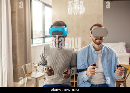 Zwei multikulturelle fröhliche Menschen, lässig gekleidet, Spaß mit neuen Trends Technologie, Sitzen, Seite, Seite mit VR-Brille und Controller in Han. Stockfoto
