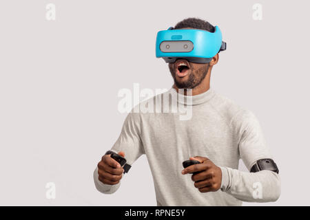 Dunkelhäutige Glücklich, aufgeregt, der Mann mit der kurzen Stoppeln, pullower in Weiß gekleidet, trägt blaue mobile virtual reality Headset halten sich an den Händen, als ob er Stockfoto