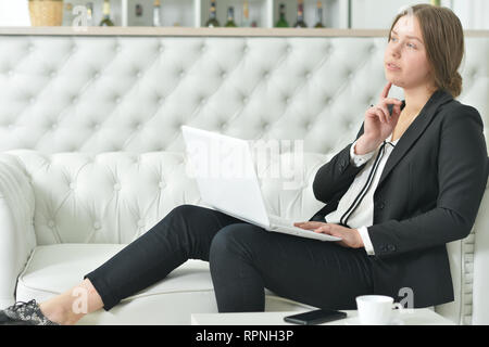 Nachdenklich jugendlich Mädchen tragen Gesellschaftsmode mit Laptop beim Sitzen auf dem Sofa Stockfoto