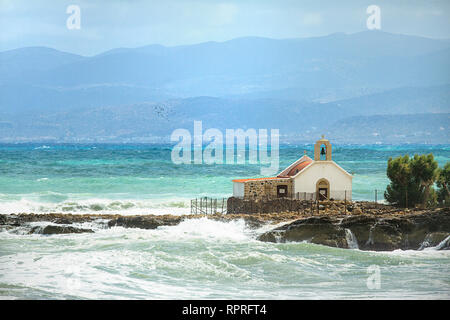 Grobe Mittelmeer, Agios Nikolaos Kirche auf einer Klippe in Hersonissos, Berge im Hintergrund. Windiges Wetter. Scharen von Vögeln. Insel Kreta, Griechenland Stockfoto