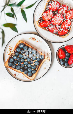Blick von oben auf die süße Toasts mit Nutella, Beeren, abgestürzte Mandel- und Kokos Chips auf weißer Tisch. Flach, Frühstück Konzept. Stockfoto