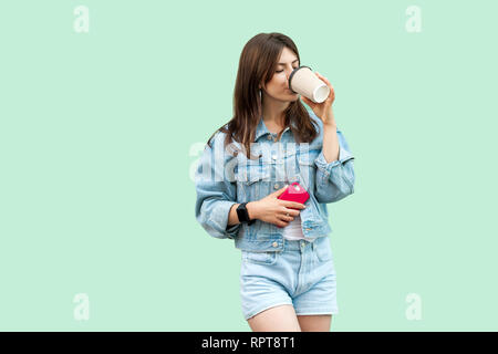 Porträt der schöne Junge brünette Frau in casual Denim Style stehen und Holding einweg Becher, Trinken und Ausruhen und Genießen. indoor Studio Stockfoto