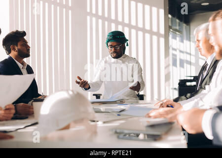 Arabische Investor in Weiß nationalen candura und gutra auf Kopf diskutieren Unternehmen finanzielle Strategie mit seinem multirucial männlichen Managern während der Konferenz ein Stockfoto