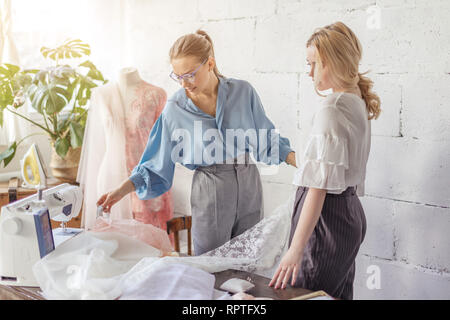 Junge hübsche blonde Frau wählt Stoff für ihr Hochzeitskleid und weiblichen professionellen Schneider in stilvoller blauer Bluse Ihr in hilft Stockfoto