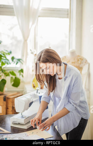 Innen- schuss der kaukasischen jungen Frau in Blau casual aufgeknöpfte Hemd nimmt Nadeln für die Modellierung Kleid von Arbeitstisch in der Werkstatt. Stockfoto