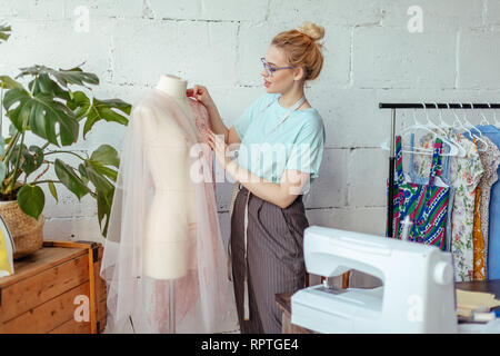 Geschickte Fashion Designer steht in einer Schneiderin Studio in der Nähe der Puppe und Fehlerbehebungen Spitzenstoff mit Nadeln. Schaffung oder Modellierung der Mode Stockfoto