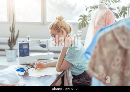 Verträumt nachdenklich Näherin mit blonden hairmaking Notizen in Nähen Studio in Rosa und Mint Töne, einen Arbeitsplan für die nächste Woche Stockfoto