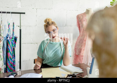 Attraktive junge Couturier in Brillen sitzen auf dem Desktop mit Skizzen, in die Kamera schauen und lächeln, während der Arbeit im Atelier im alten Stil Stockfoto
