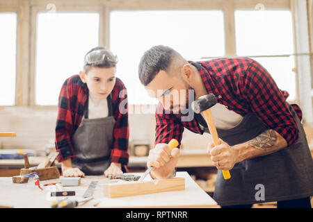 Bärtige vorschäler Mann in eine Schutzbrille und eine Schürze arbeitet mit Meißel in eine Schreinerei. Tischler mit einem Meißel und sein Sohn beobachten den Prozess. Stockfoto