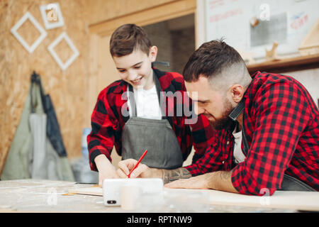 Konzentrierte sich Handwerker und seine kleine Anhänger Junge sind Messung von Holz mit einem Lineal und Bleistift in die Werkstatt. Erwachsene Mann vergleicht die korrekte Markup einschalten lässt Stockfoto