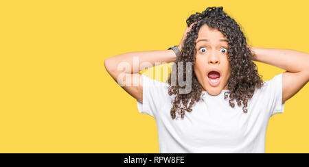 Junge schöne Frau mit lockigem Haar tragen weiße t-shirt, verrückt und Angst mit den Händen am Kopf, Angst und überrascht von Schock mit offenen Mund Stockfoto