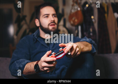 Porträt eines jungen Mannes Spielen von Videospielen, die auf dem Rand seines Sitzes auf der Wohnzimmer Couch. Fokus auf Video Game Controller. Stockfoto